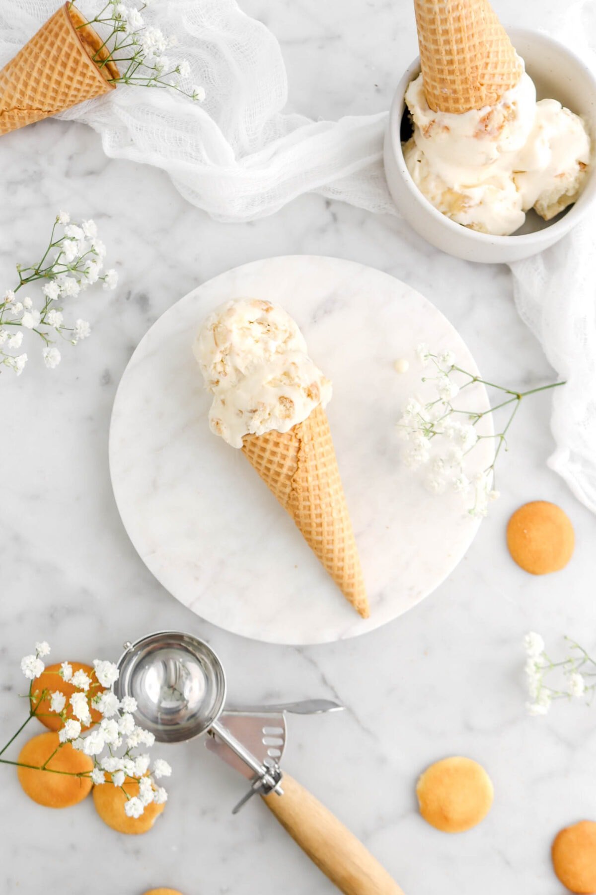 overhead shot of two scoops of banana pudding ice cream in sugar cone on marble plate with bowl full of ice cream beside, flowers and vanilla wafers around, and an antique ice cream scoop