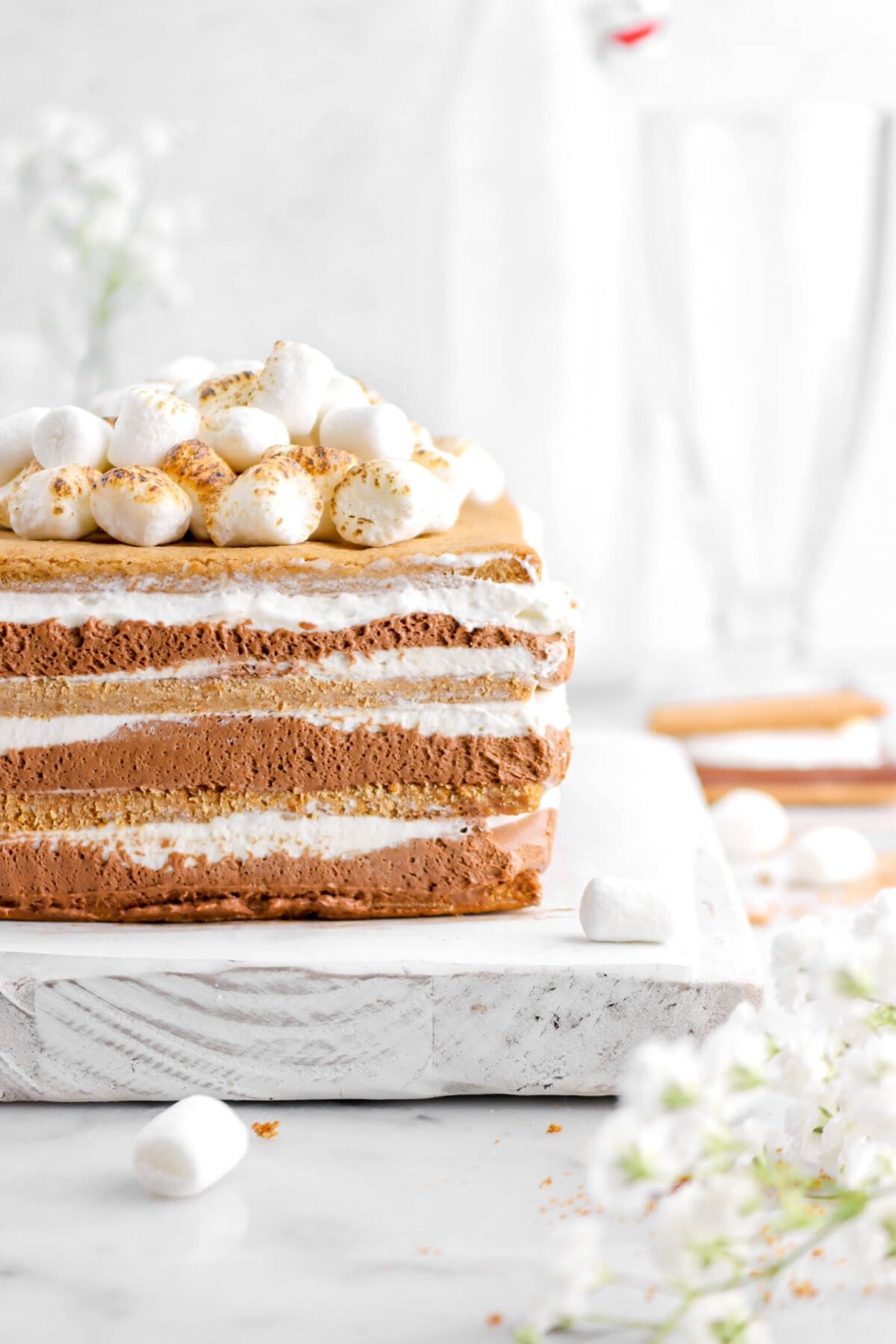 close up of icebox cake with marshmallows on top, sitting on a white wood board, with flowers beside and mini marshmallows around
