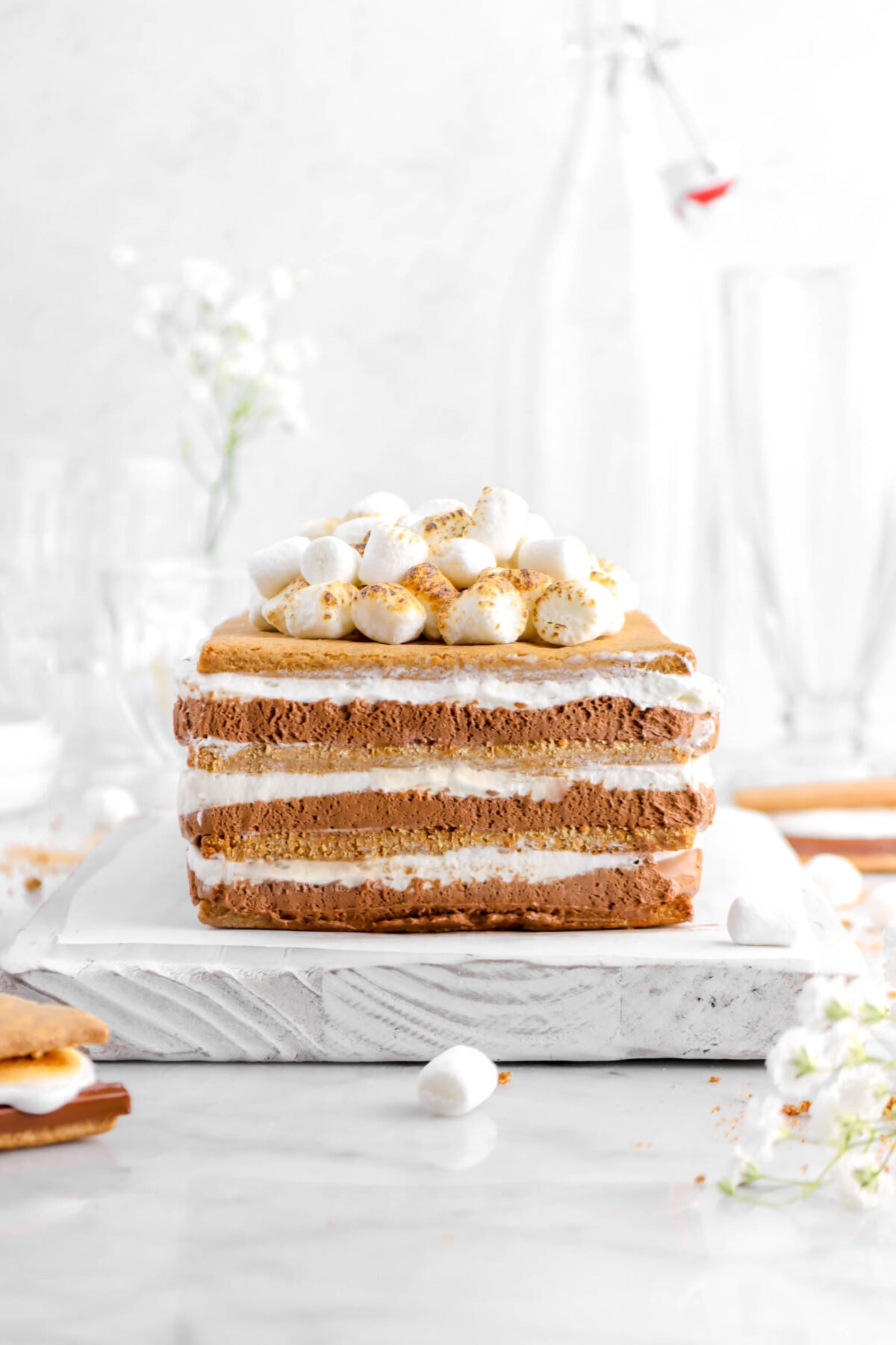 front shot of s'mores icebox cake on white wood board with mini marshmallows on top and flowers behind