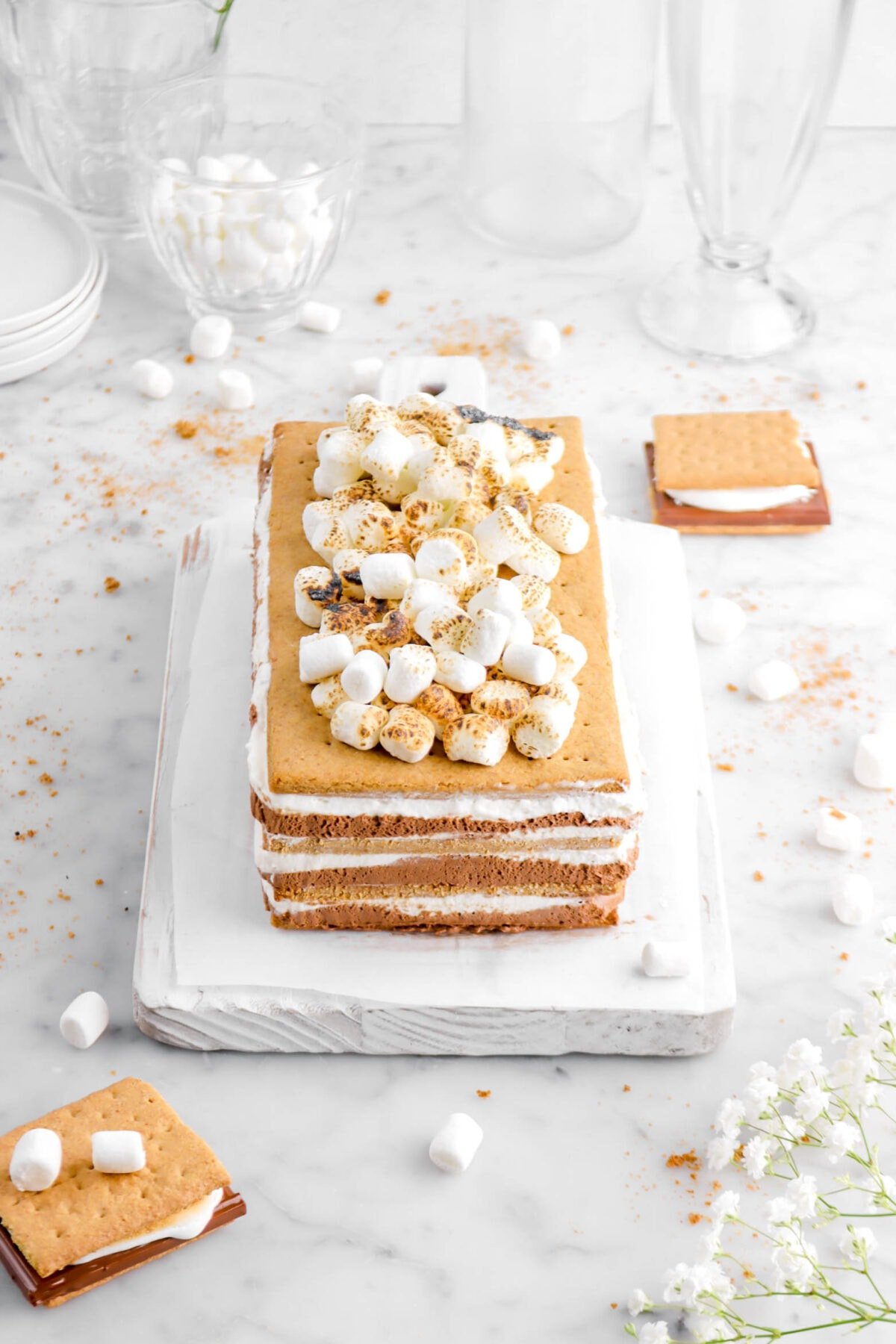 close up angled photo of icebox cake on wood serving board on a piece of parchment paper with two s'mores beside and mini marshmallows around