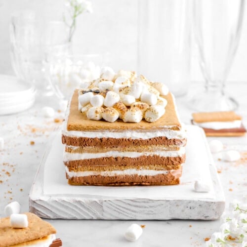 s'mores icebox cake with two s'mores beside, mini marshmallows toasted on top, empty glasses behind, with graham cracker crumbs and mini marshmallows scattered around on marble surface