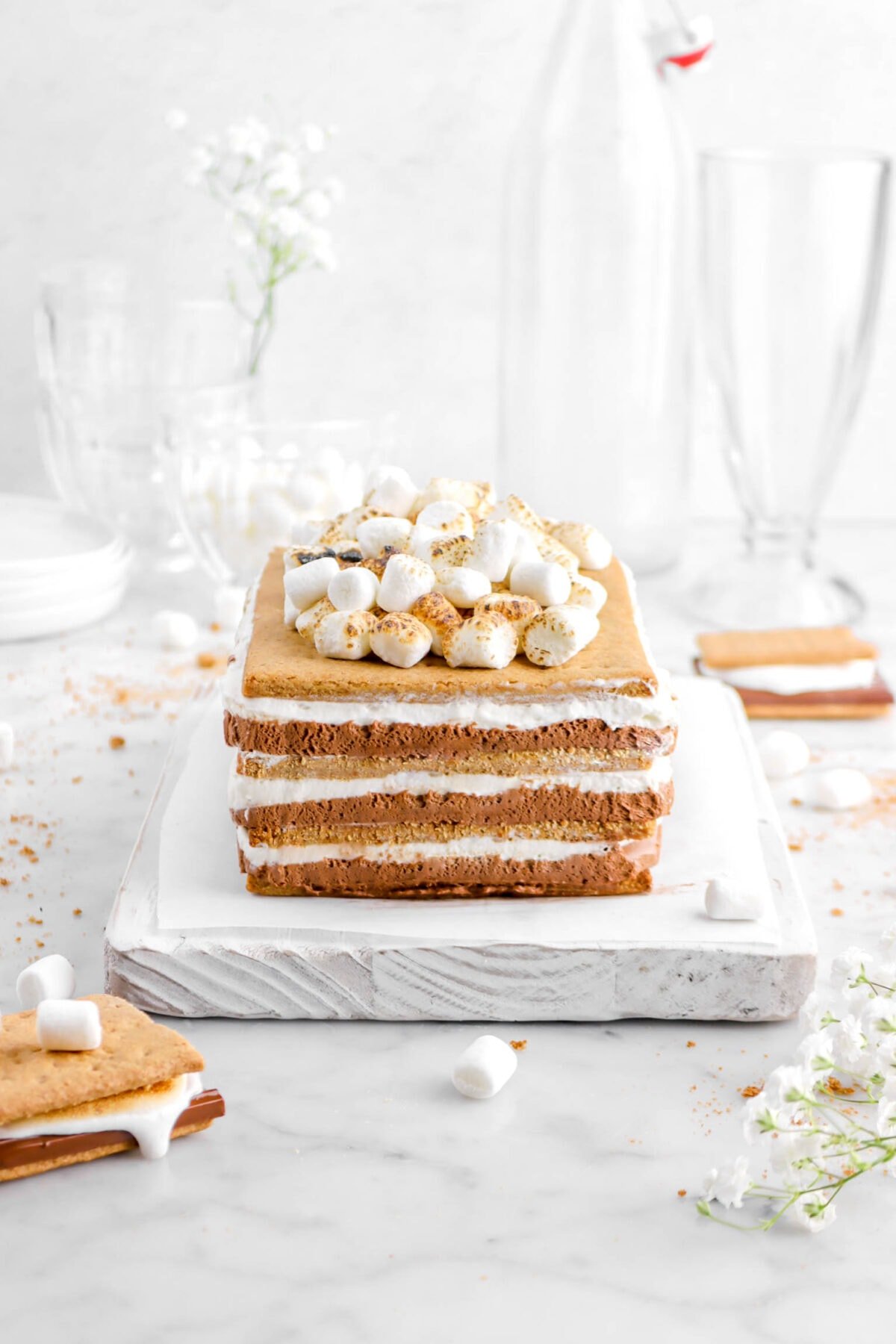 s'mores icebox cake with two s'mores beside, mini marshmallows toasted on top, empty glasses behind, with graham cracker crumbs and mini marshmallows scattered around on marble surface