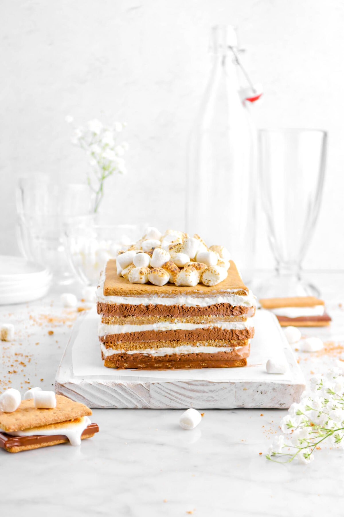 icebox cake with toasted marshmallows on top on wood board with flowers, two'smores, and graham cracker crumbs around with glass of mini marshmallows behind