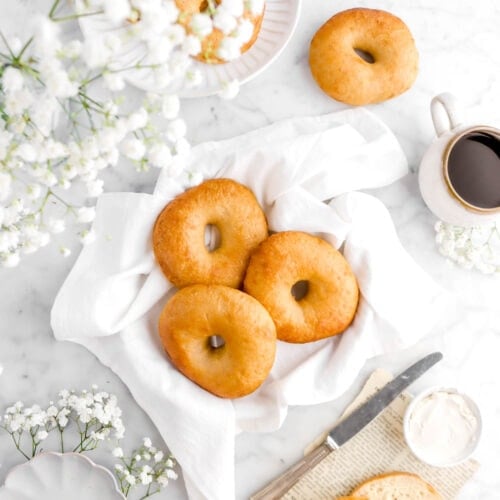 bagels on white napkin with more bagels around, one cut open beside, with a knife and cream cheese beside, a cup of coffee, and flowers around