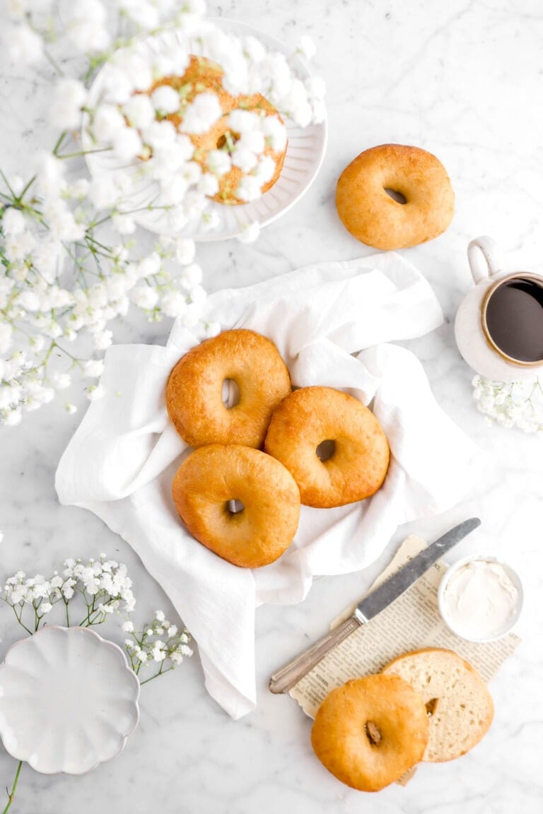 bagels on white napkin with more bagels around, one cut open beside, with a knife and cream cheese beside, a cup of coffee, and flowers around