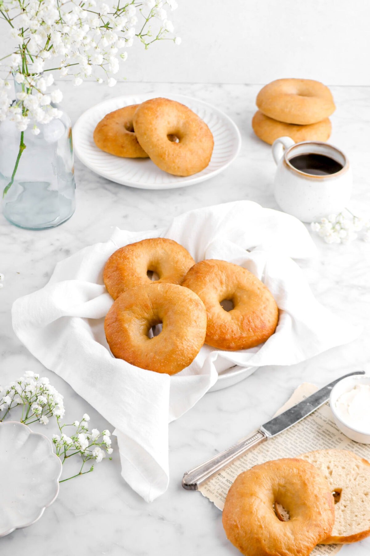 angled shot of three bagels in pie plate with white napkin, four bagels behind with a glass vase of flowers, and cup of coffee
