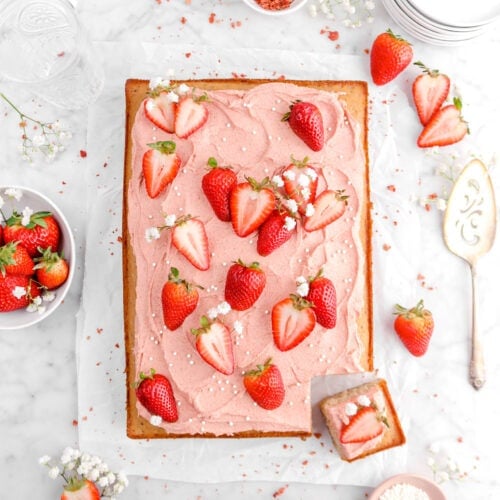 overhead shot of sheet cake with strawberry frosting, fresh strawberries, pearl sugar, and little white flowers on top, fresh strawberries around, white flowers, cake serve, and stack of white plates