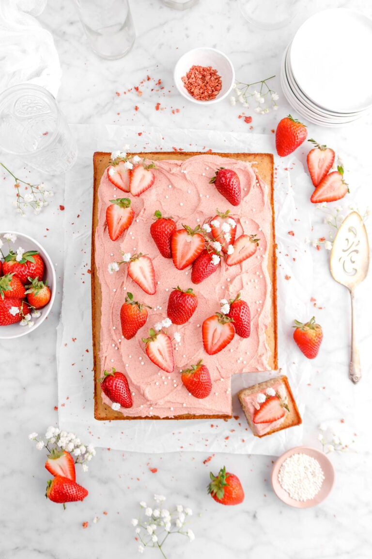 overhead shot of sheet cake with strawberry frosting, fresh strawberries, pearl sugar, and little white flowers on top, fresh strawberries around, white flowers, cake serve, and stack of white plates