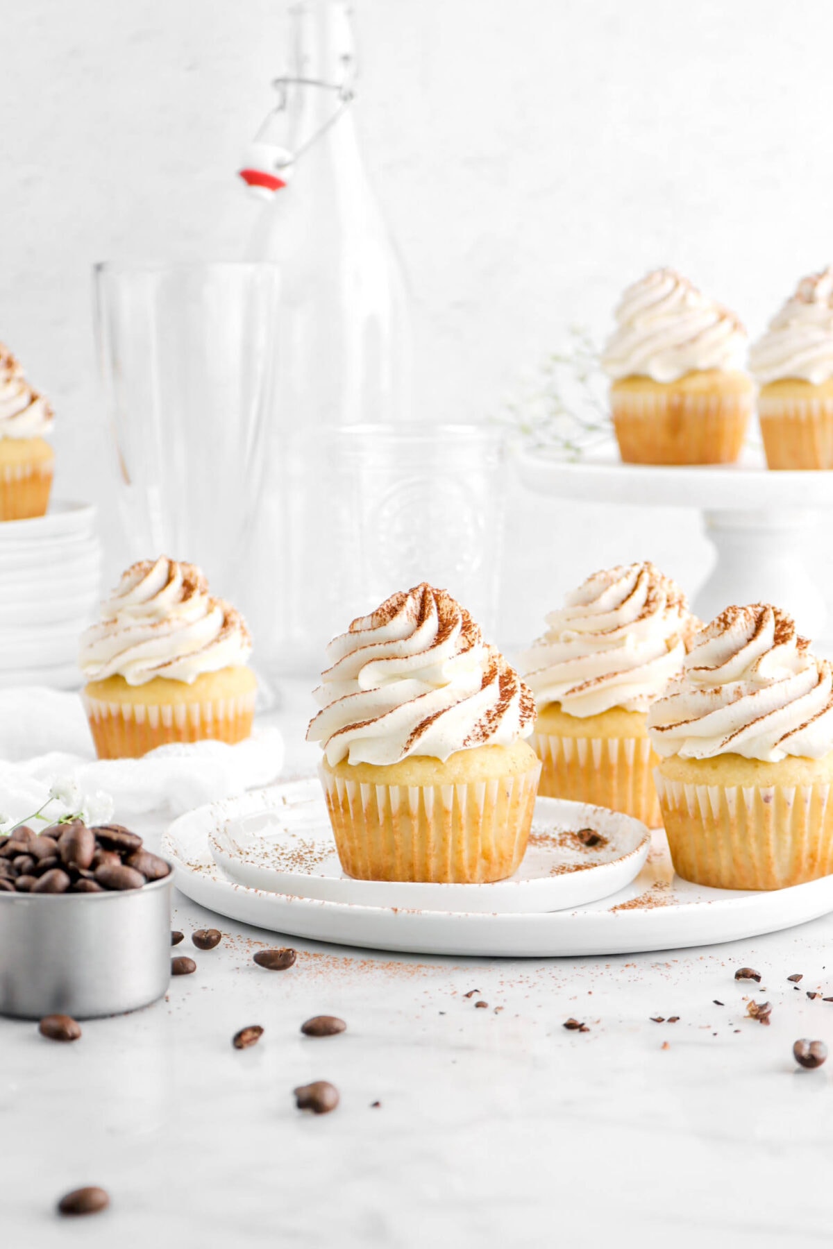 front shot of tiramisu cupcakes on white plates with coffee beans scattered around, a cake plate behind with empty glasses