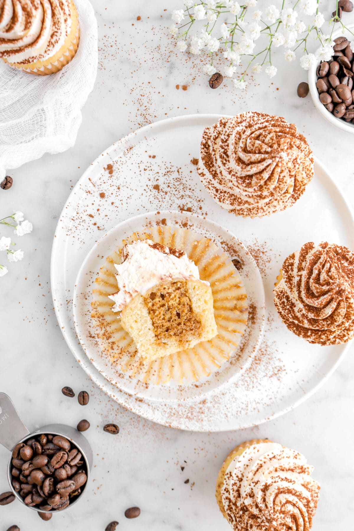 overhead shot of cupcake sliced in half on cupcake paper, with two white plates underneath, cocoa powder, coffee beans, and more cucakes around