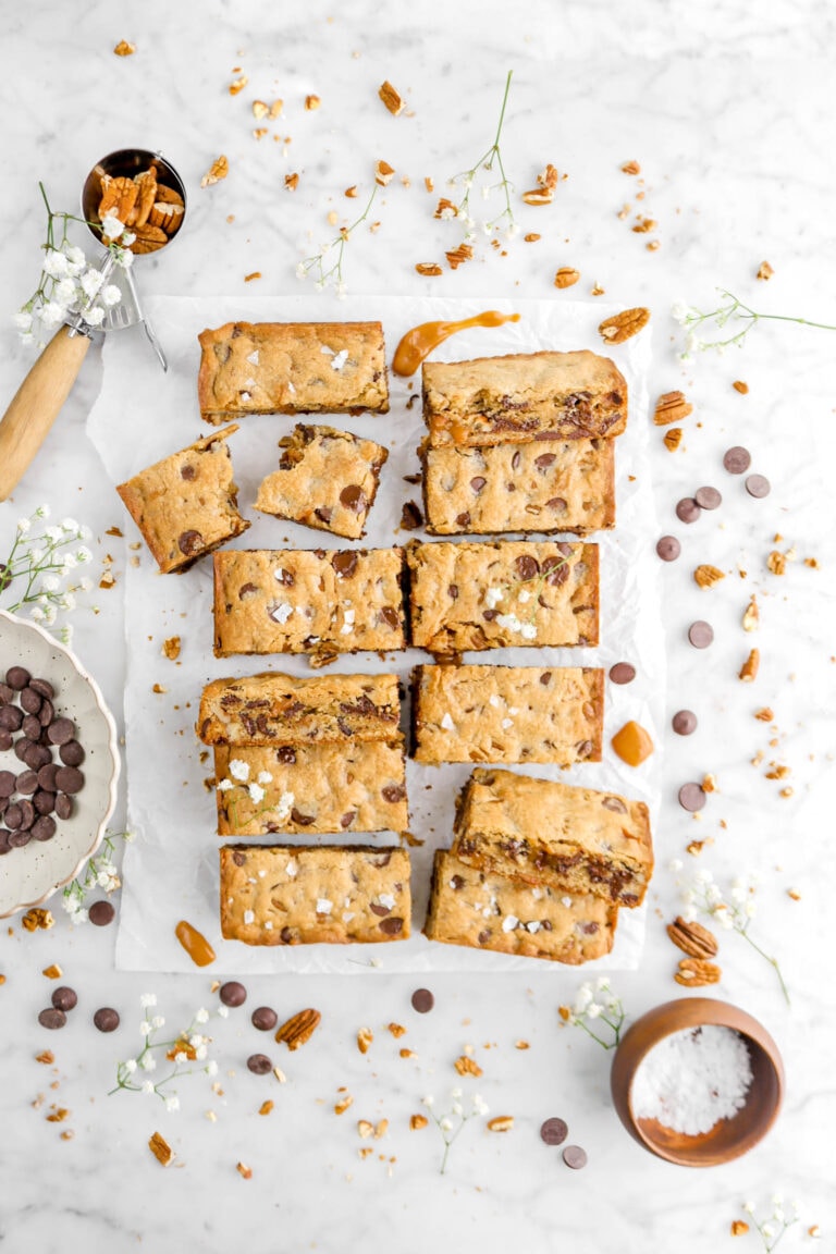 turtle cookie bars on parchment paper with ice cream scoop full of pecans, caramel pieces and chocolate chips around, with small white flowers and wooden bowl of salt beside