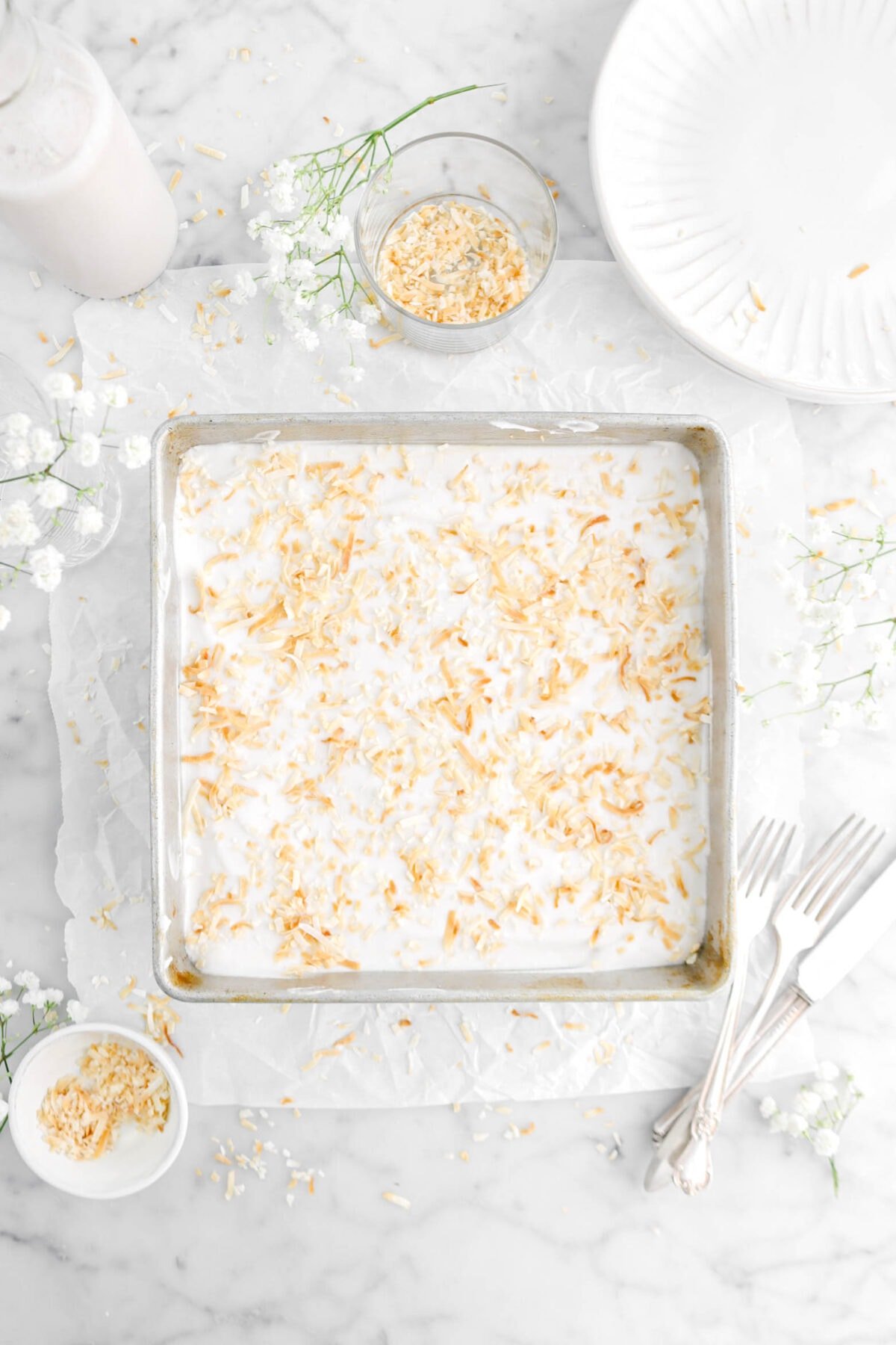 cake with whipped cream and toasted coconut on top in square cake pan on parchment paper with toasted coconut around, flowers, forks, and plates on marble surface
