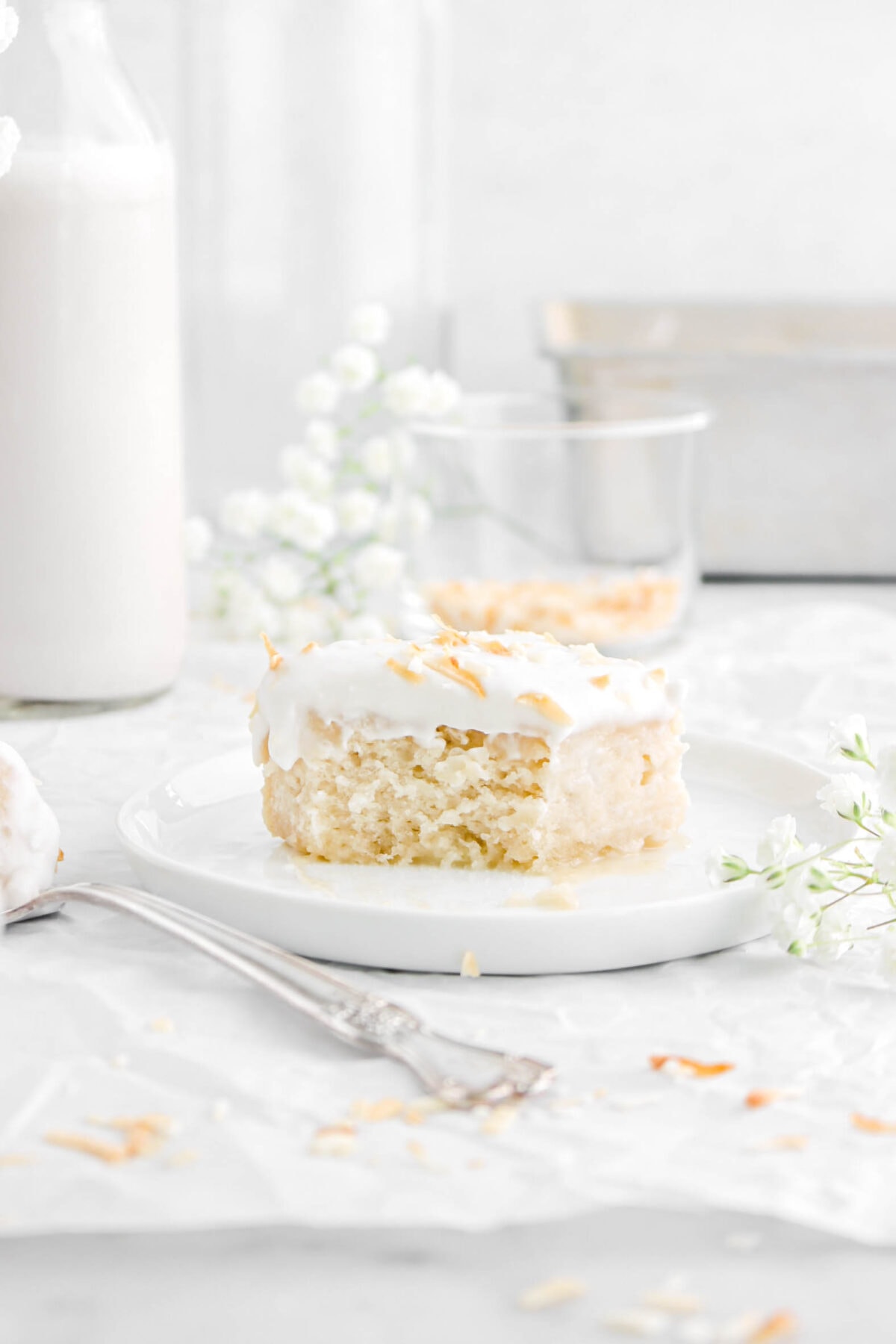 bite missing from slice of coconut cake on white plate with glass of milk and flowers behind