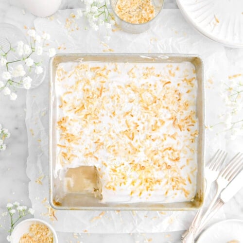 coconut tres leches cake in square pan with square slice on white plate beisde, toasted coconut scattered around, two forks, and white flowers around