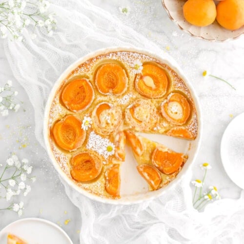 sliced apricot clafoutis in tart pan with chamomile flowers on top, on a white cheese cloth and bowl of apricots beside