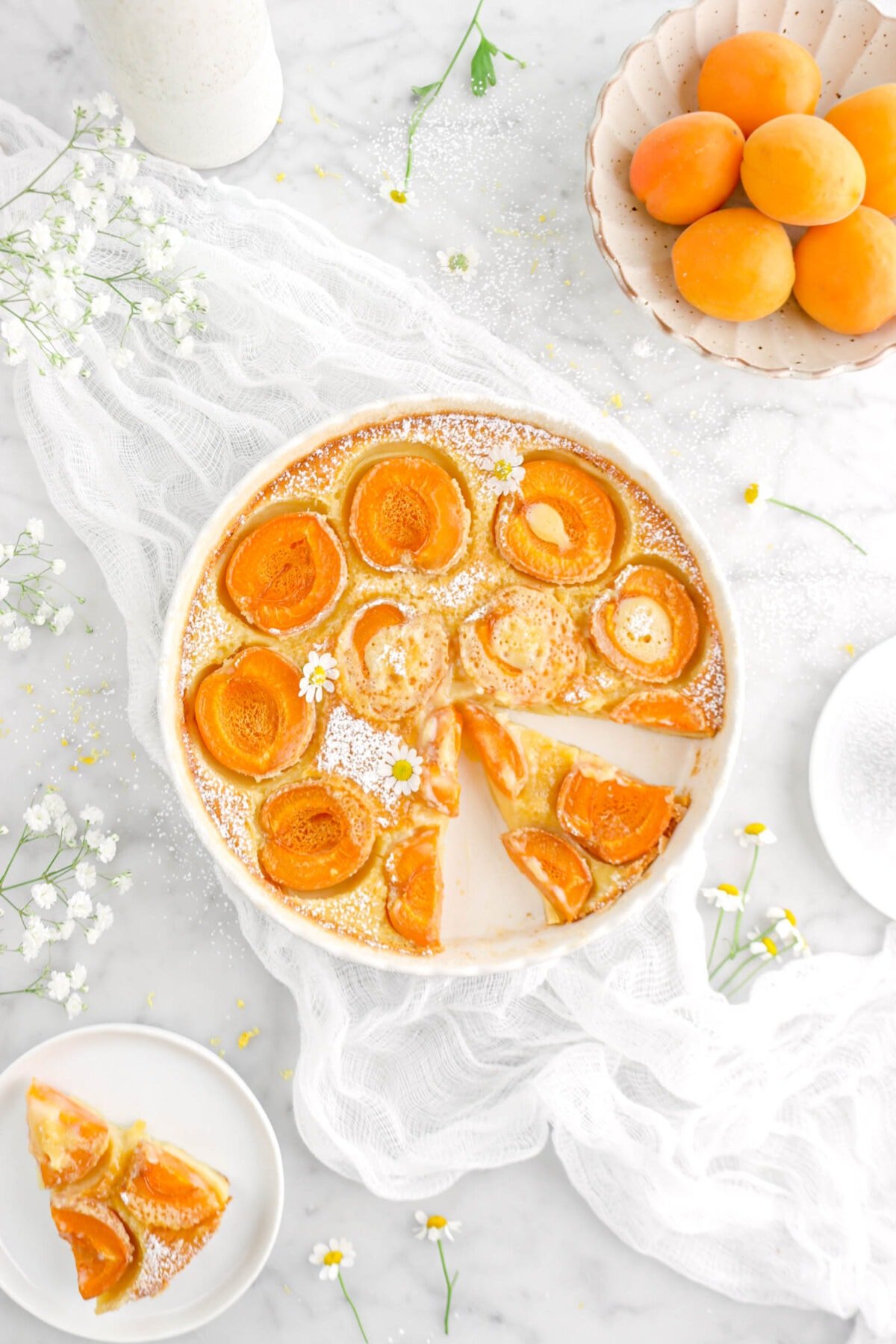 sliced apricot clafoutis in tart pan with chamomile flowers on top, on a white cheese cloth and bowl of apricots beside
