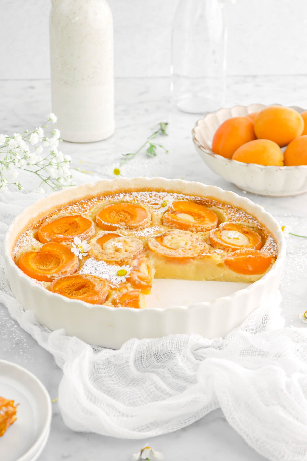 front shot of apricot clafoutis with slices missing on white cheesecloth with bowl of apricots and vase behind
