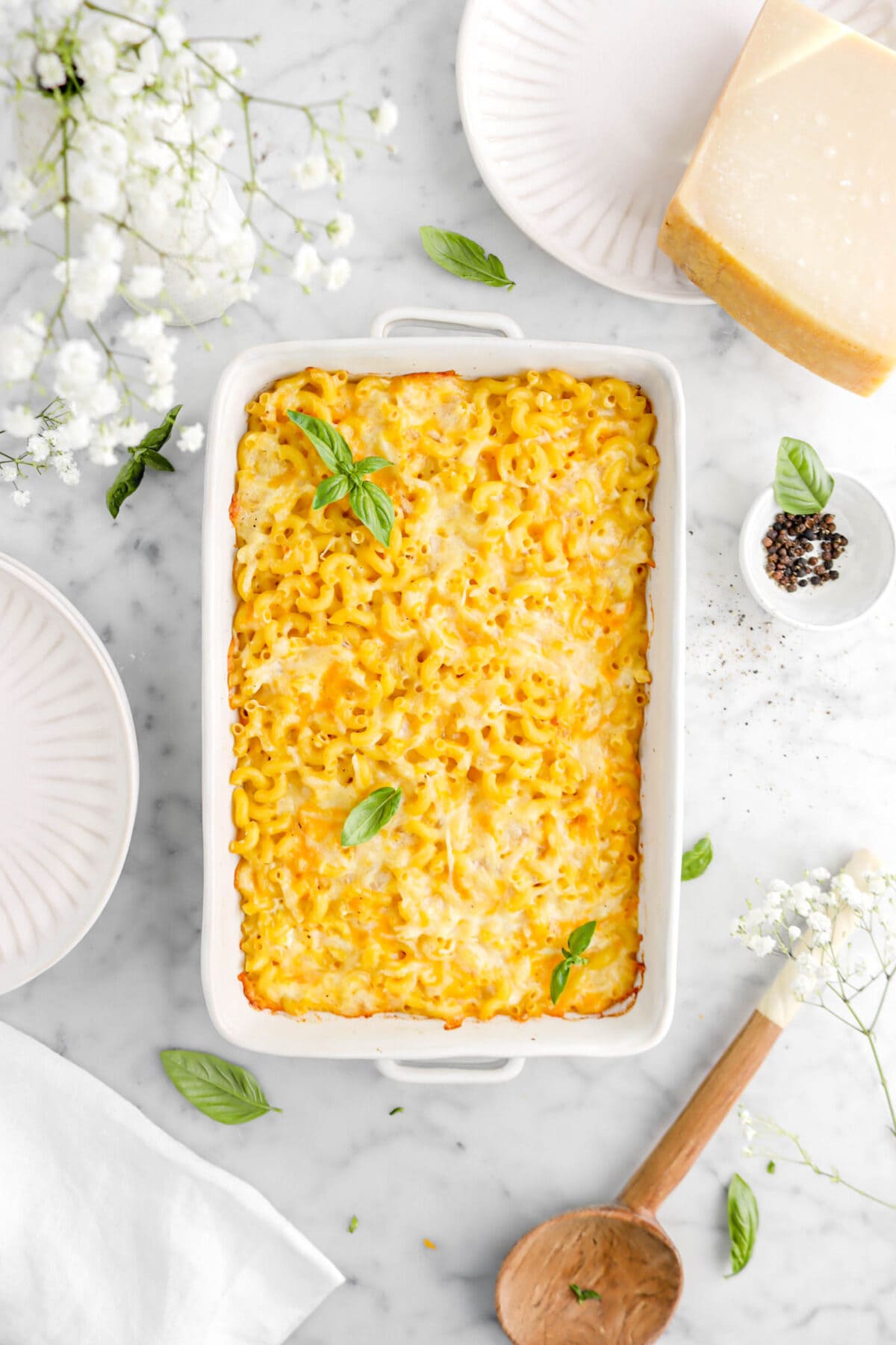 overhead shot of mac and cheese in white casserole with flowers and basil leaves around on marble surface, with plates beside and wooden spoon