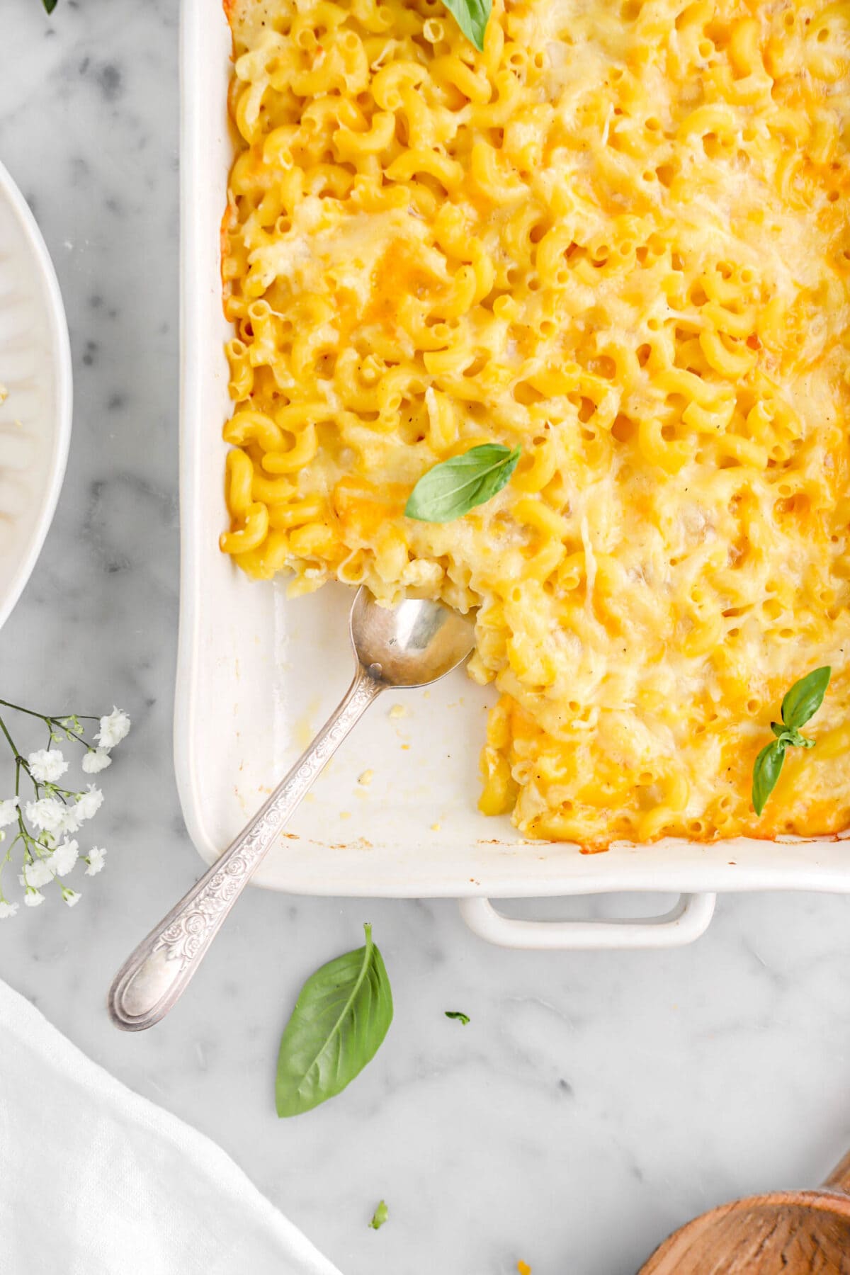 baked macaroni and cheese with spoon dug into it on marble surface with basil leaves and white flowers around