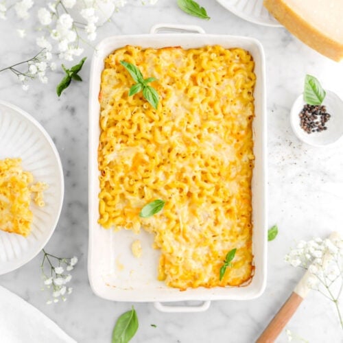 overhead shot of baked mac and cheese in white casserole with slice on plate beside, flowers and basil leaves around, a small bowl of peppercorns, and block of parmesan cheese beside,