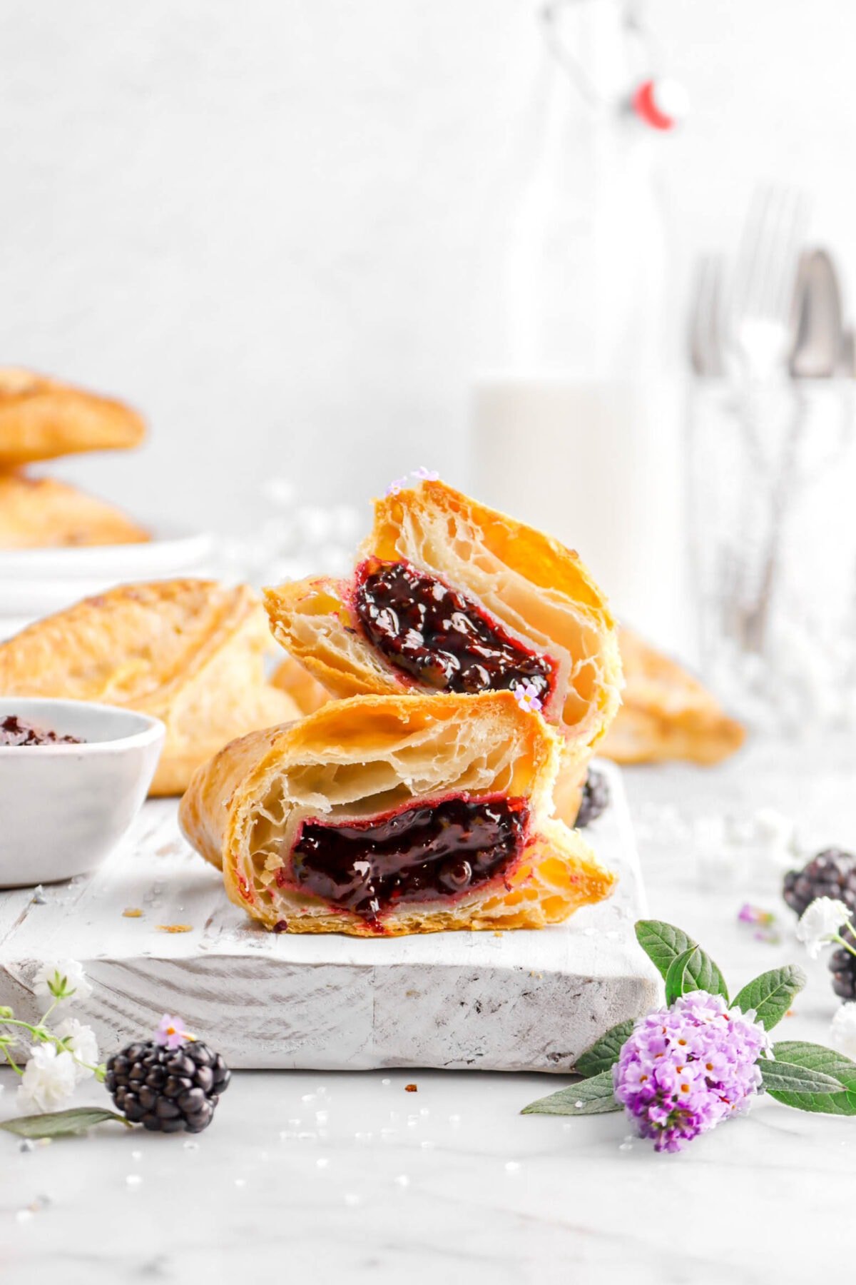 close up of sliced blackberry turnover on white wood board with fresh blackberries, purple flowers, and bowl of jam beside, with more turnovers and a glass of milk behind