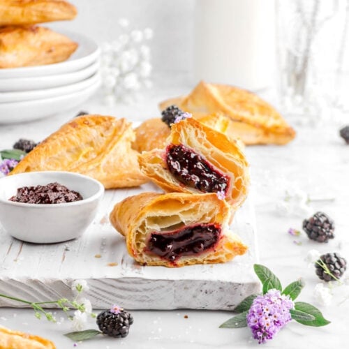 two blackberry turnover halves stacked on a white wood board with four more turnovers behind, a bowl of jam beside, with fresh berries, purple and white flowers around, glass of milk, and glass of antique utensils behind