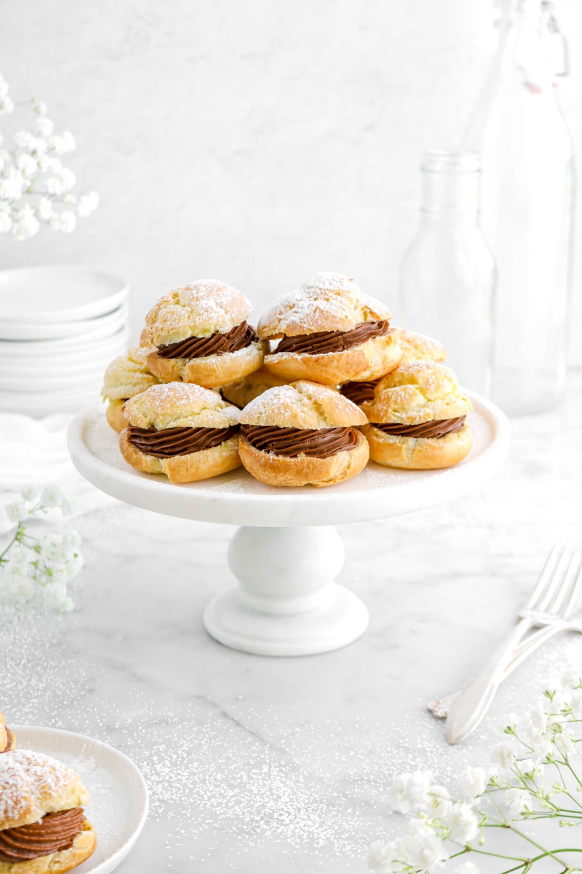 angled close up of cream puffs with flowers around, two forks beside, a stack of plates behind, and empty glasses