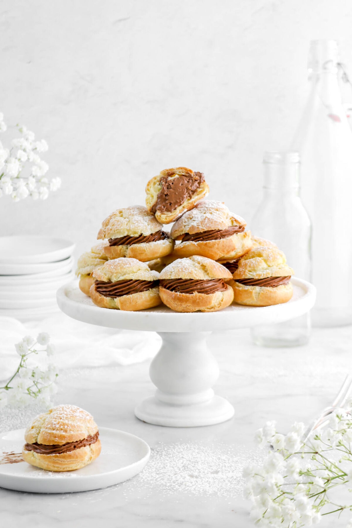nine chocolate cream puffs stacked with top one cut open on cake plate with flowers around