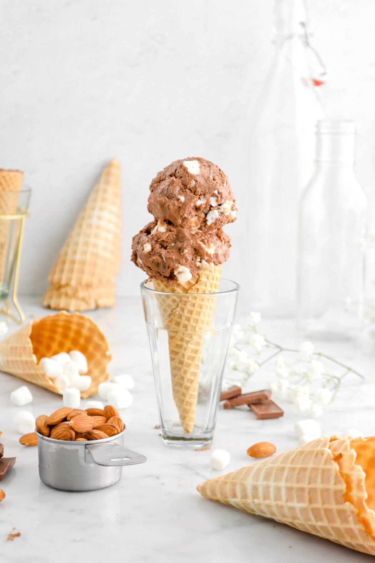 ice cream in glass standing up with cones around, mini marshmallows, almonds, and chocolate pieces on marble surface