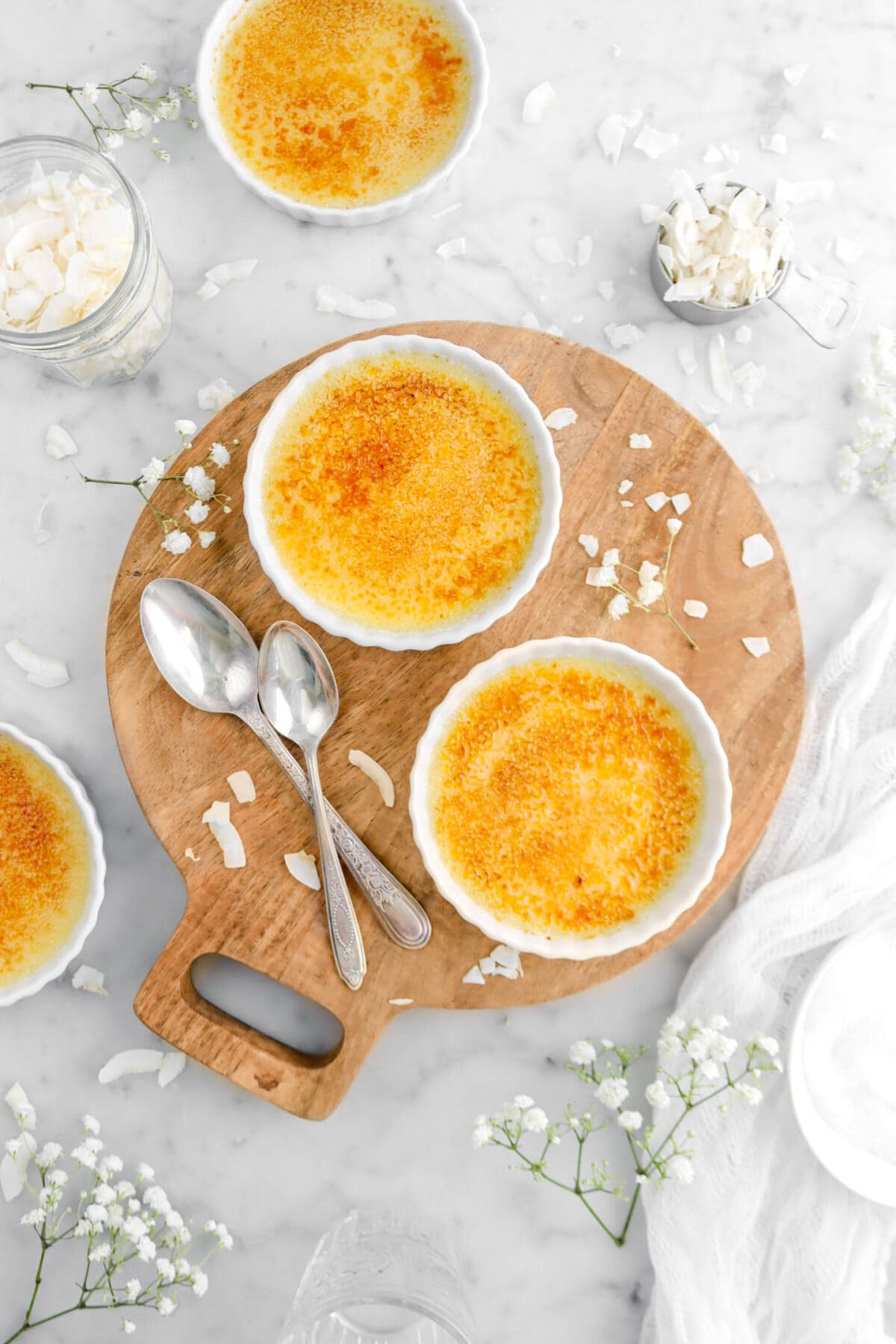 two creme brulee's on round wood board with two spoons beside, coconut flakes around, and flowers beside