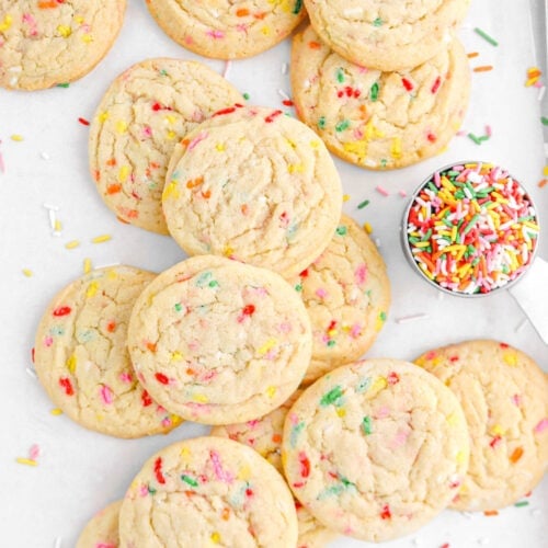 overhead shot of stacked funfetti cookies on parchment paper with measuring cup full of sprinkles