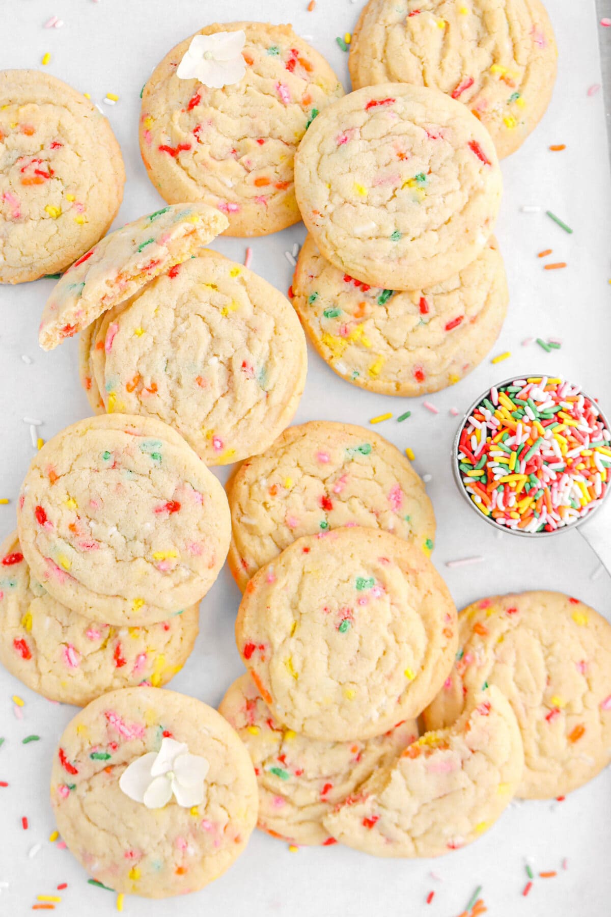 funfetti cookies piled on parchment paper with one cookie broken in half, two white hydrangea flowers, and a measuring cup of rainbow sprinkles beside