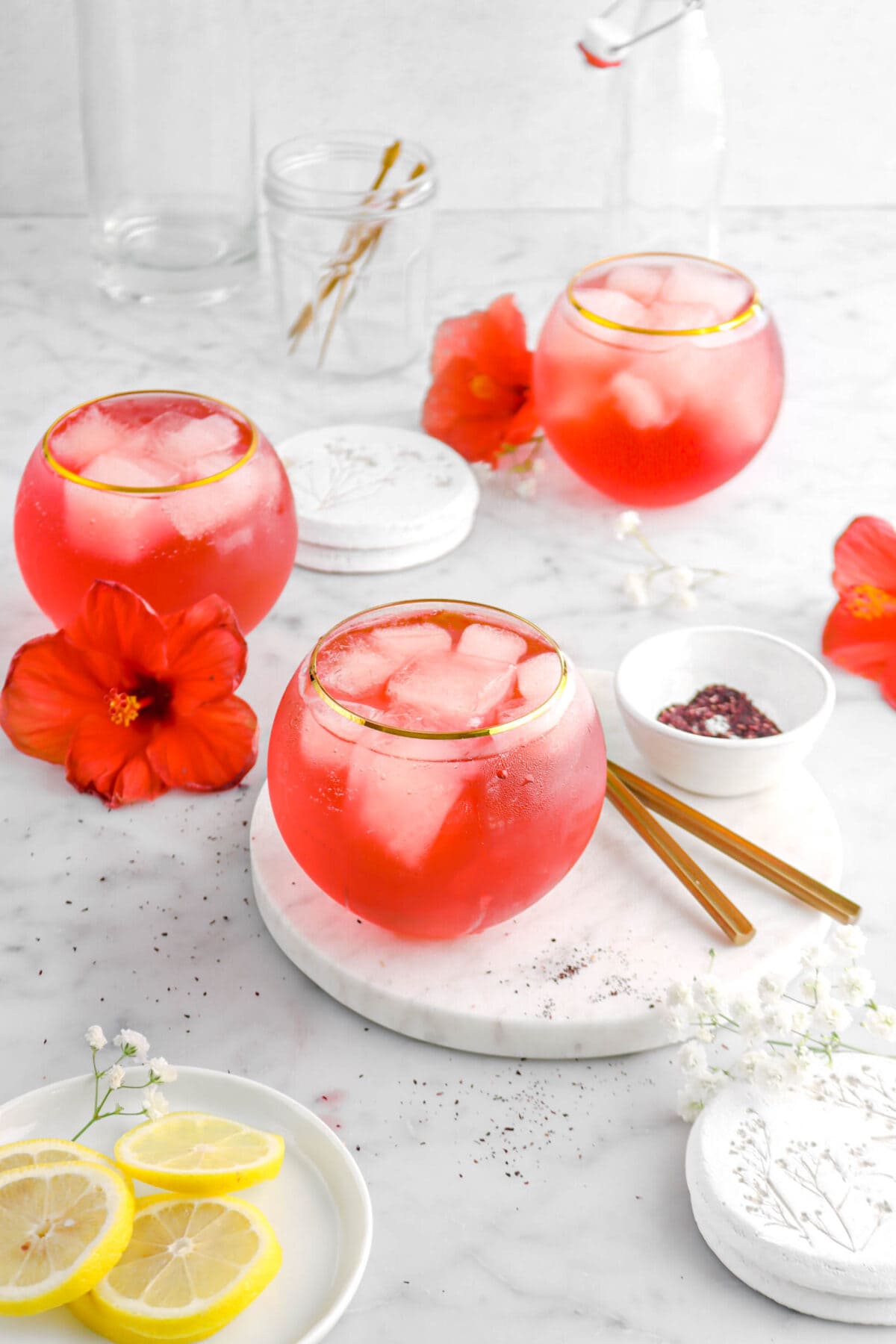 angled shot of three glasses of hibiscus cocktail on marble surface with hibiscus flowers, white flowers, and and lemon slices beside