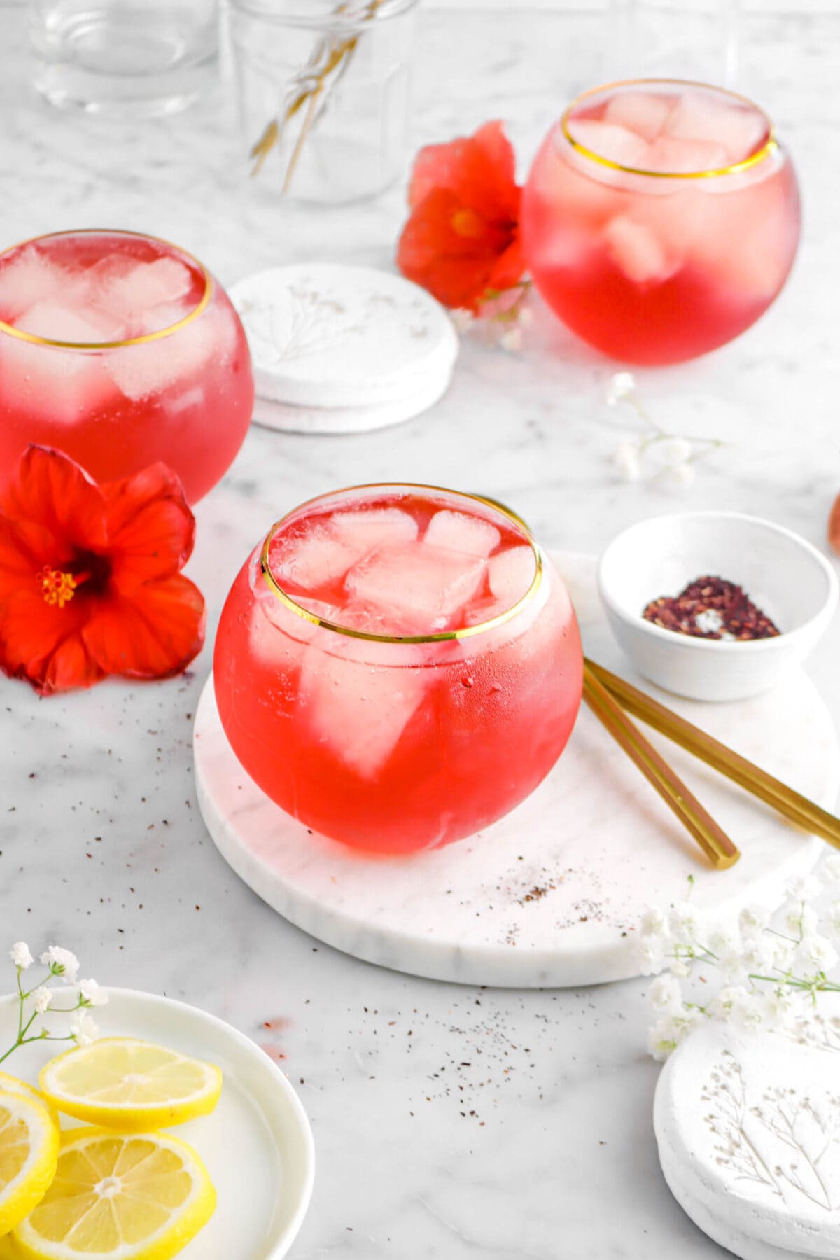 angled shot of hibiscus cocktail on marble surface with bowl of dried hibiscus beside, two flowers, and two more drinks behind