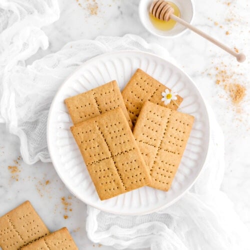 four graham crackers on white plate with white flower on top, with two more graham crackers beside, a white cheese cloth, crumbs around, and a bowl of honey on marble surface
