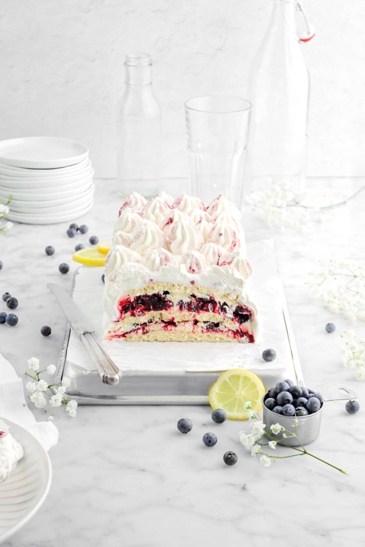 pulled back and angled photo of lemon blueberry icebox cake on sheet pan with knife beside, stack of plates behind, and empty glasses