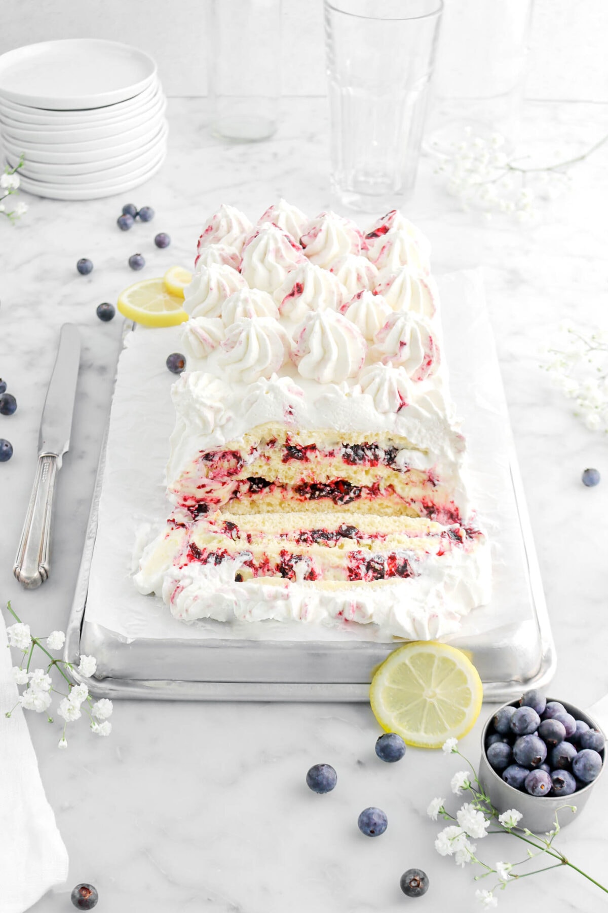 angled photo of lemon icebox cake with slice laying in front, a slice of lemon and blueberries around, with a knife and flowers beside