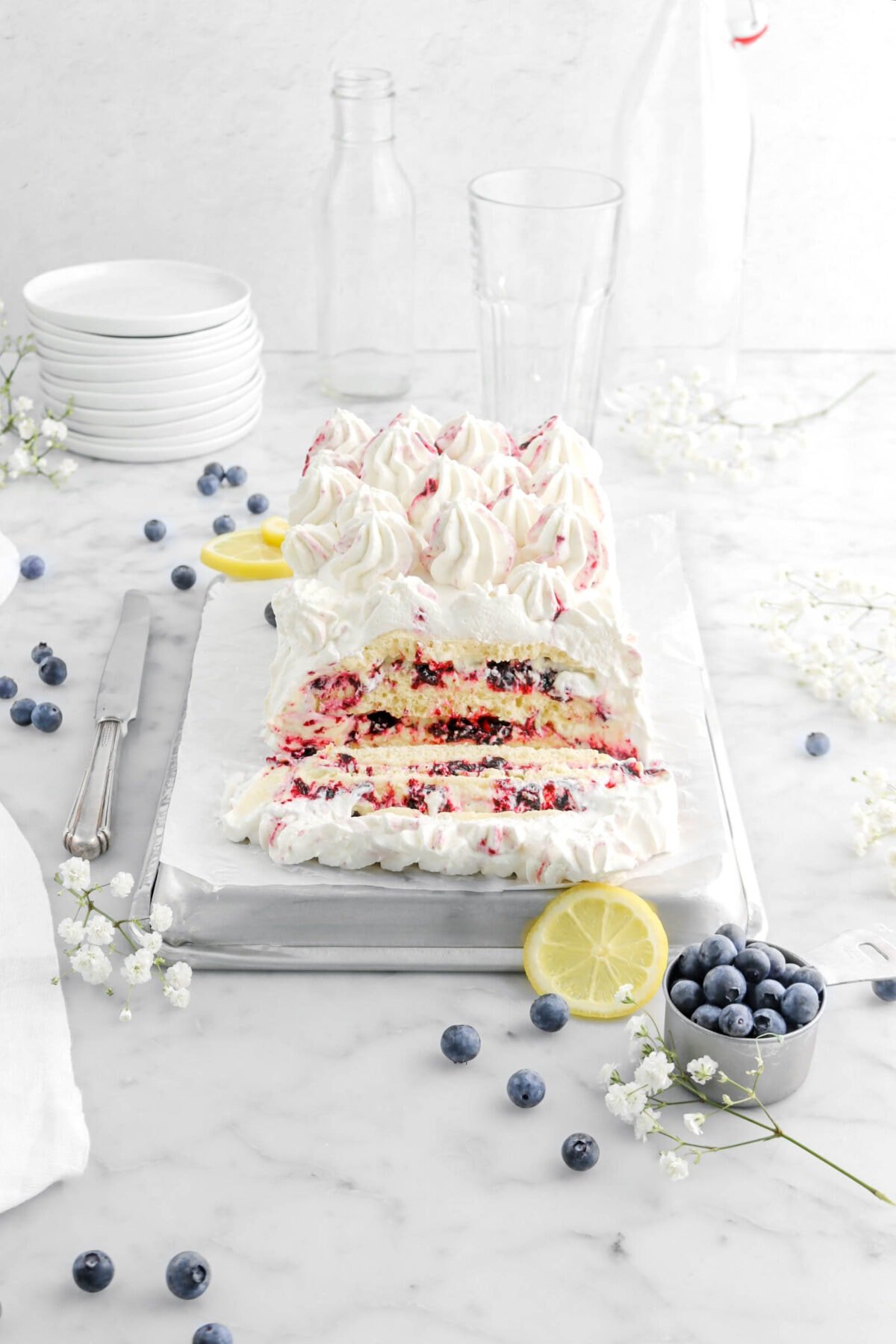 lemon blueberry icebox cake on upside down sheet pan with slice laying in front, a knife beside, blueberries, lemon slices, and flowers around, with stack of plates and empty glasses behind on marble surface