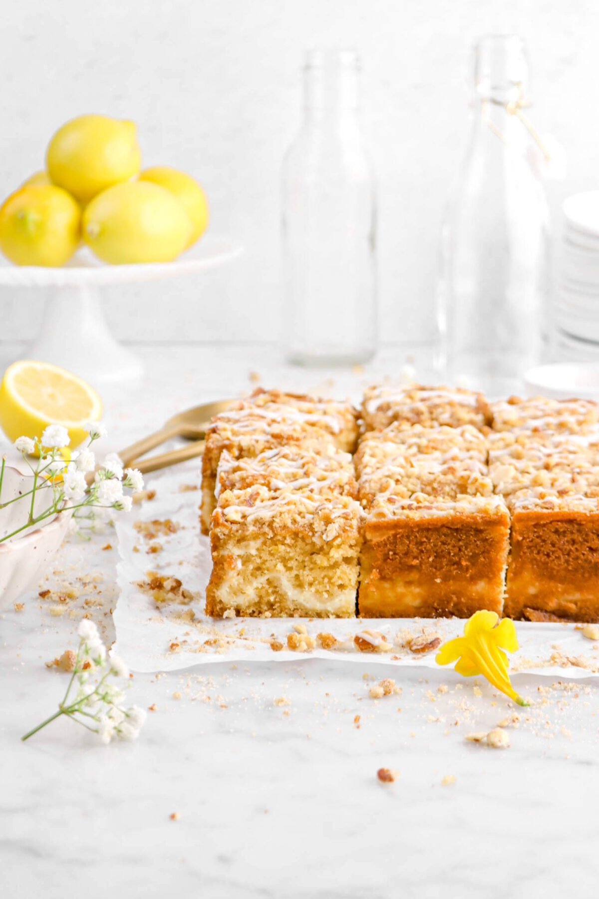 pulled back shot of lemon coffee cake on parchment paper with white and yellow flowers around, streusel scattered around, two gold spoons behind, a stack of lemons, and two empty glasses