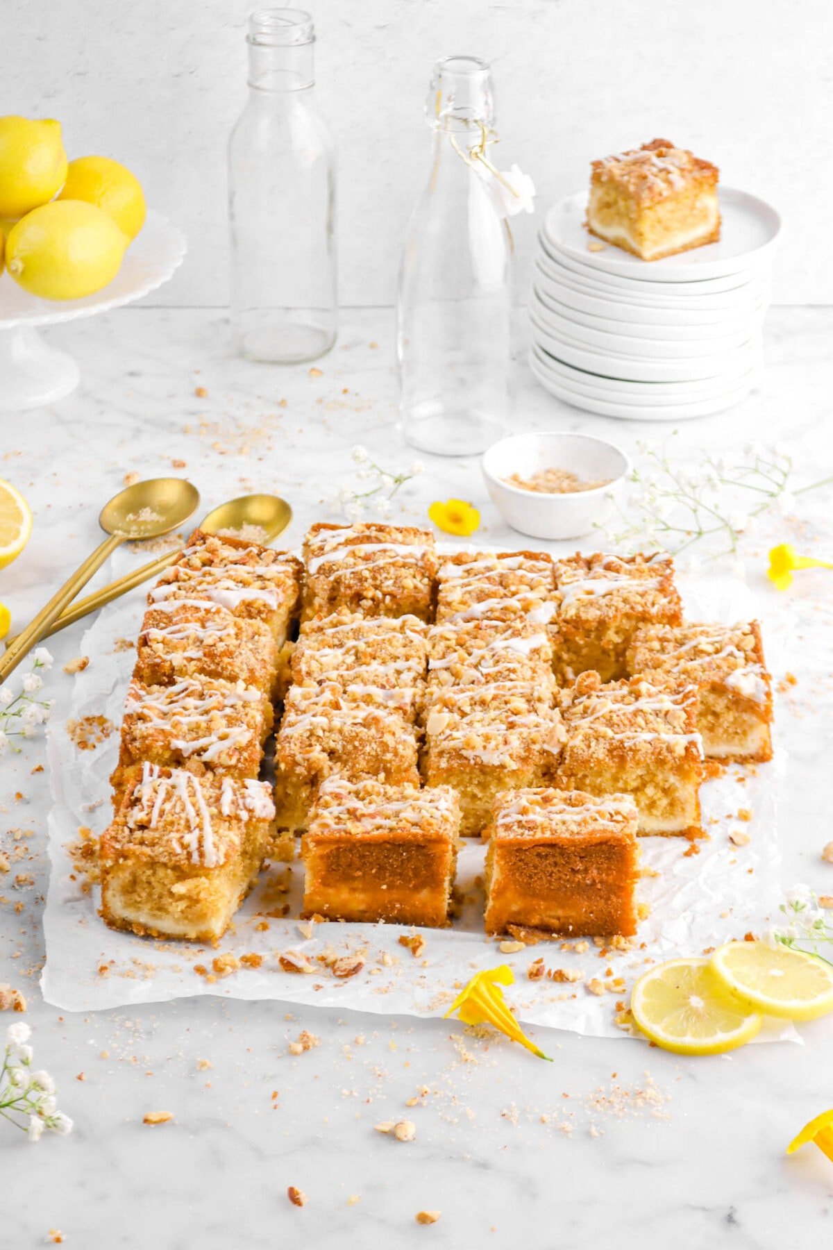 angled photo of lemon coffee cake with sliced pulled away on parchment paper, slice on stack of plates behind, with flowers around