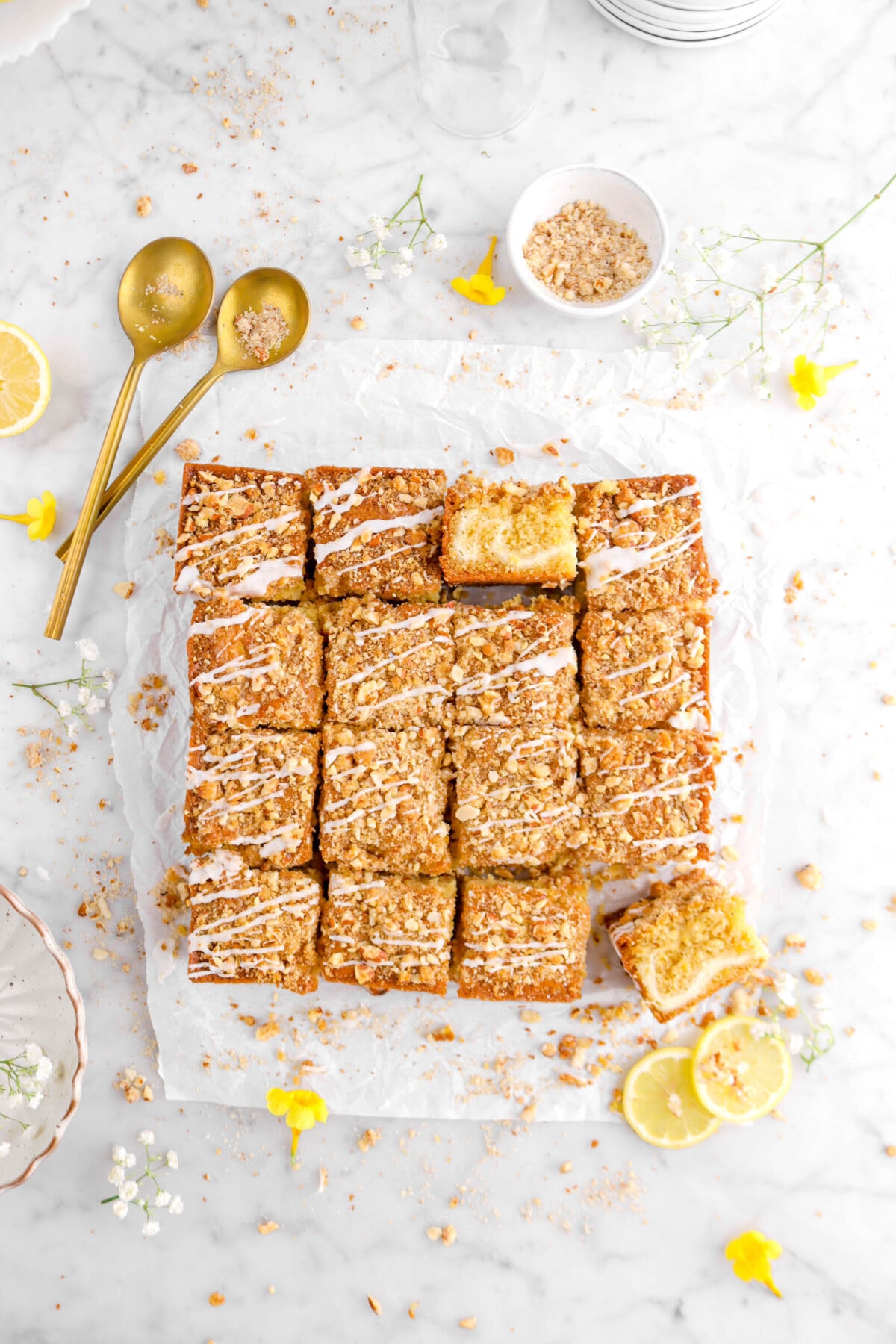overhead shot of lemon coffee cake cut into squares with two slices turned on their side, two spoons beside, two slices of lemon, a bowl of streusel, and flowers aorund