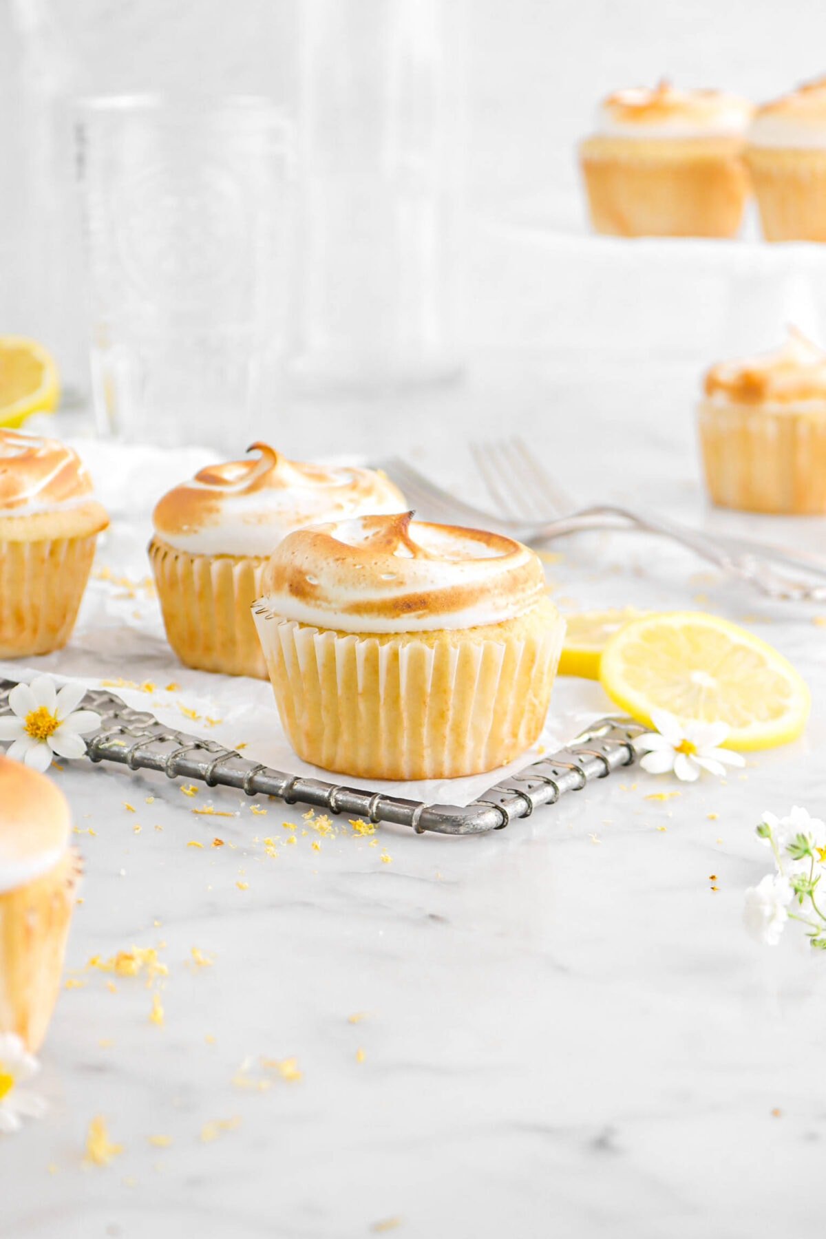 lemon meringue cupcakes on wire rack with two slices of lemon, two forks, and more cupcakes behind