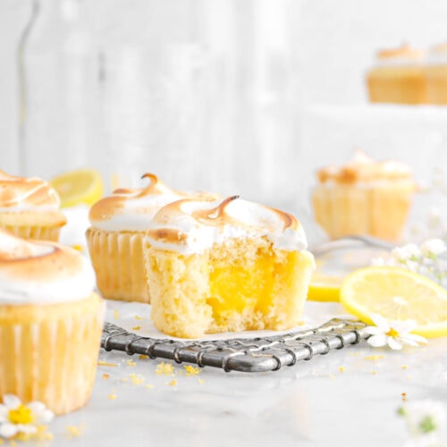 lemon meringue cupcake with bite missing on wire cooling rack, with four cupcakes around, lemon slice, flowers, and empty glasses behind on marble surface