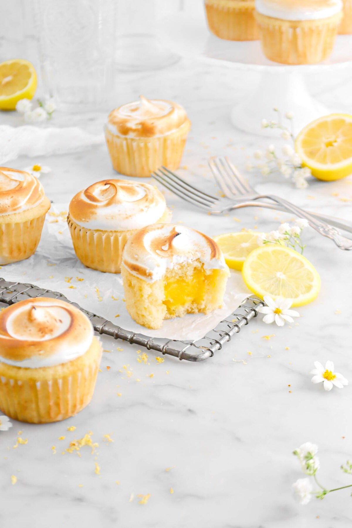 angled shot of five cupcakes with flowers and lemons around, more cupcakes on white cake stand behind