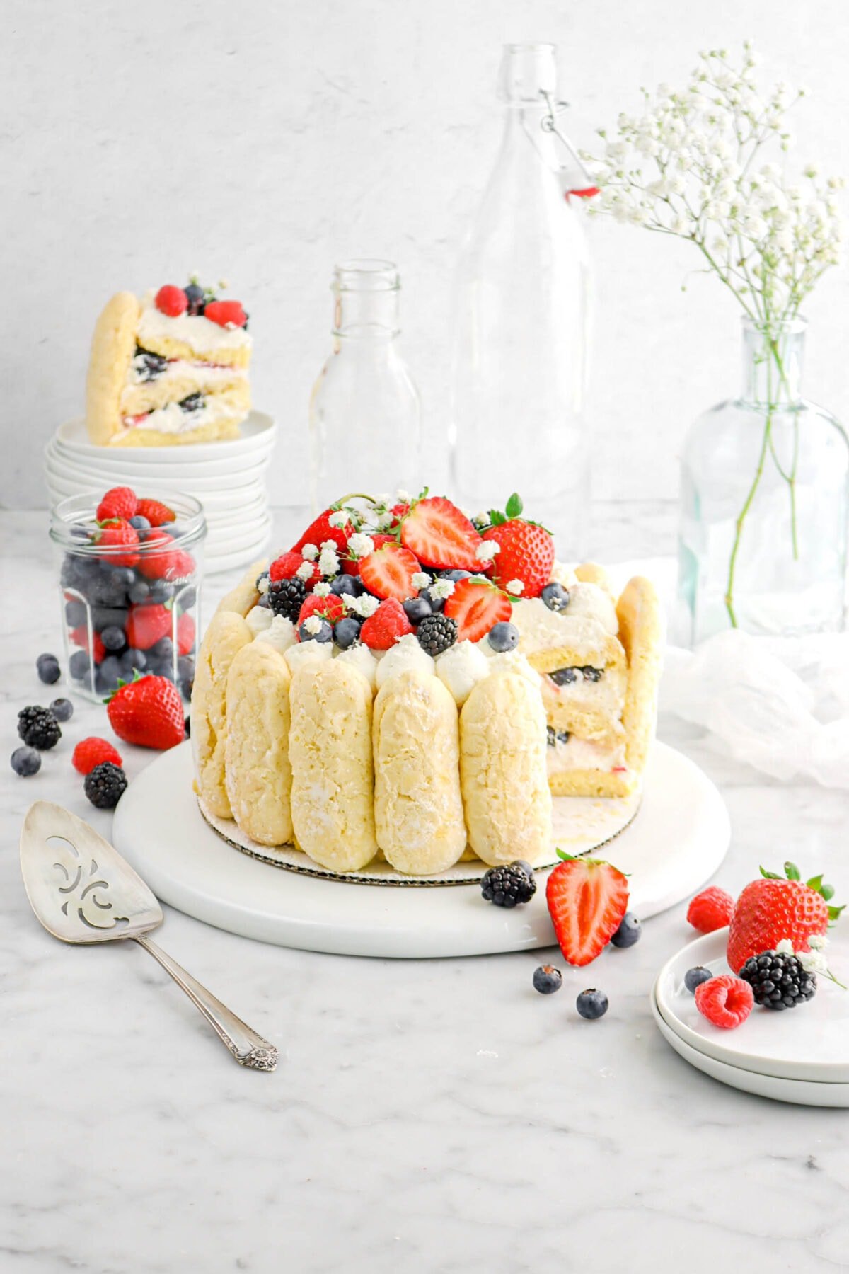 sliced tiramisu on upside down white plate with slice of tiramisu behind on stack of white plates, with a jar of mixed berries beside, with a cake knife beside on marble surface