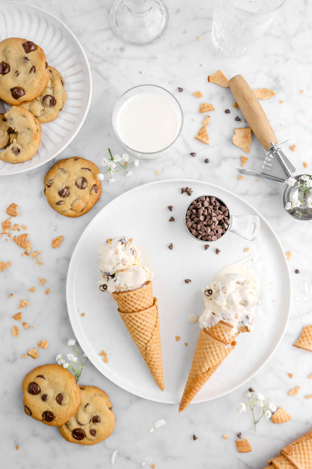 flat lay of two cones of cookie dough ice cream on white plate with chocolate chips in measuring cup beside, cookies around, and glass of milk beside