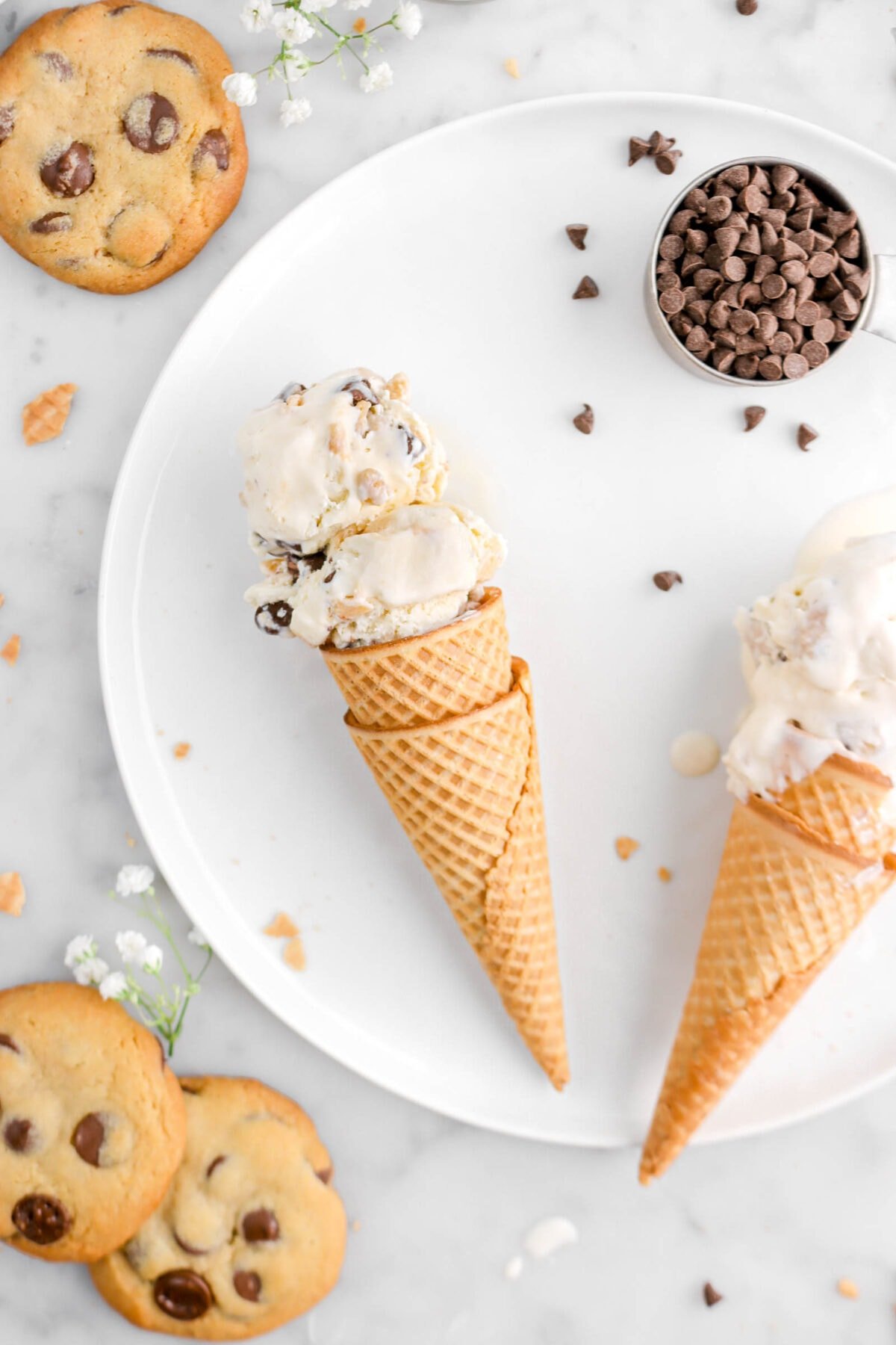 close up of double coned cookie dough ice cream laying on white plate with another cone of ice cream beside, measuring cup of mini chips, and cookies beside