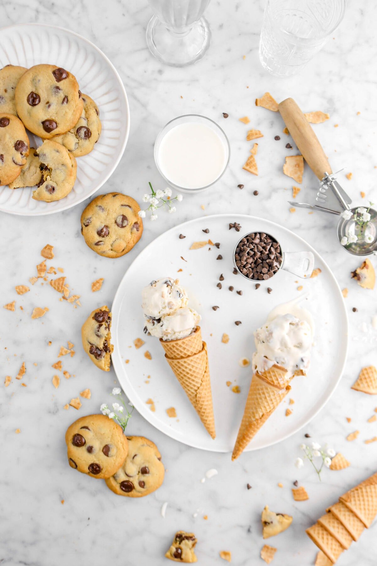 two cones of chocolate chip cookie dough ice cream on white plate with cookies, measuring cup, white flowers, mini chocolate chips, ice cream cone pieces, and ice cream scoop beside