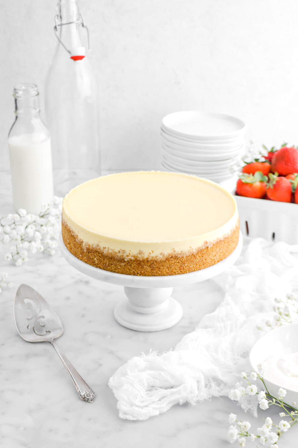 angled shot of cheesecake on marble cake plate with cake knife, and a white napkin beside, a glass of milk, stack of white plates, and basket of strawberries behind