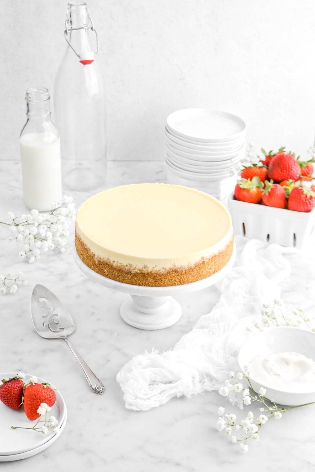 pulled back angled shot of cheesecake with plates, strawberries, flowers, cake knife, napkin, and bowl of whipped cream beside, strawberries, stack of plates, and a glass of milk behind